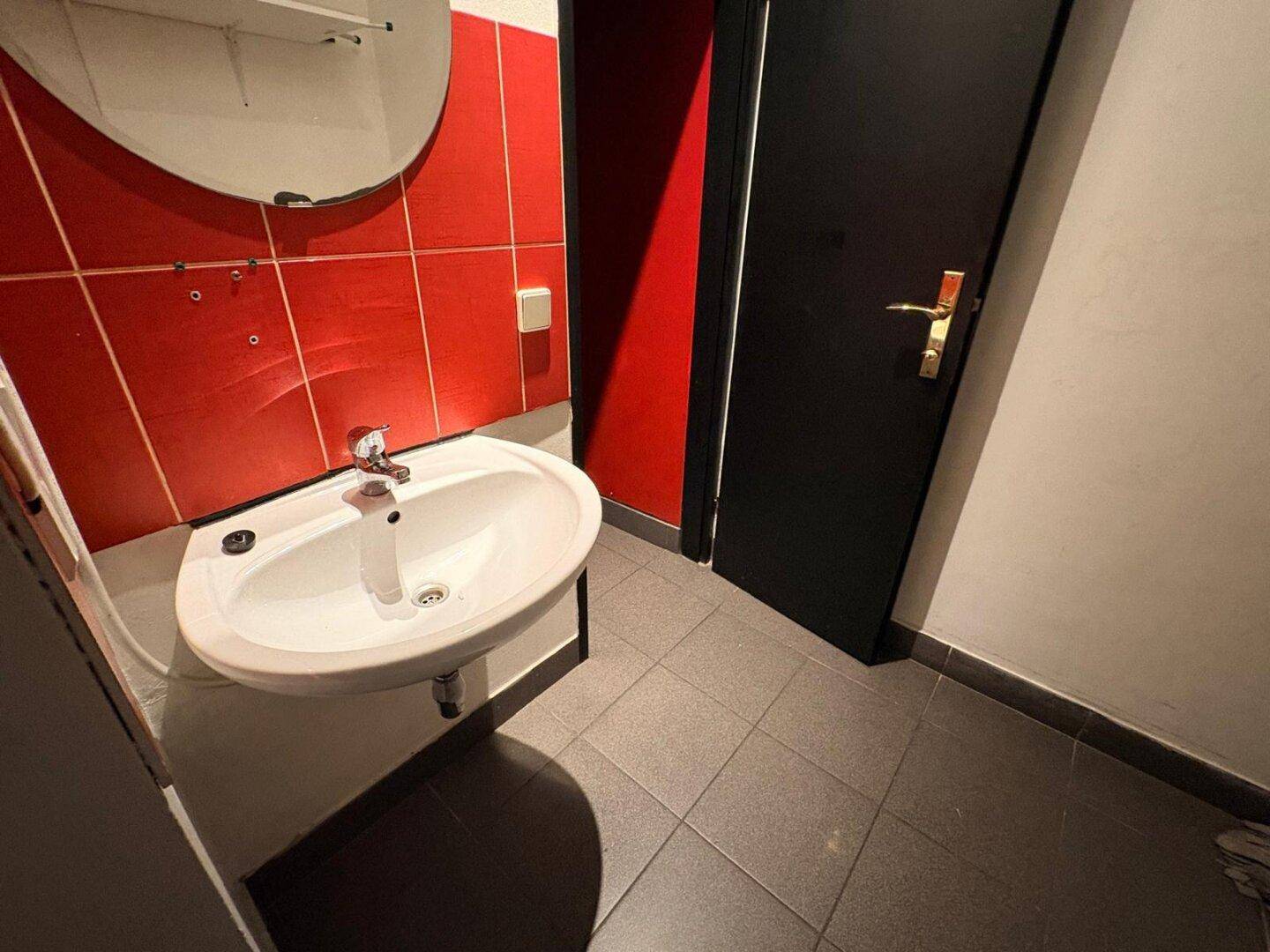 Badezimmer mit roter Wand und grauem Fliesenboden, ausgestattet mit einem Waschbecken und Spiegel.