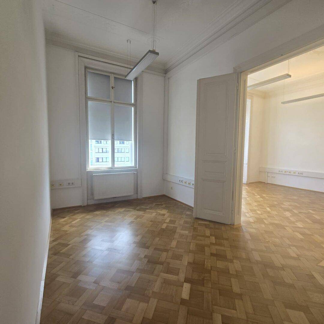 Heller Büroraum mit Fischgrätparkett, hohen Decken und einem großen Fenster, das den Raum erhellt.
