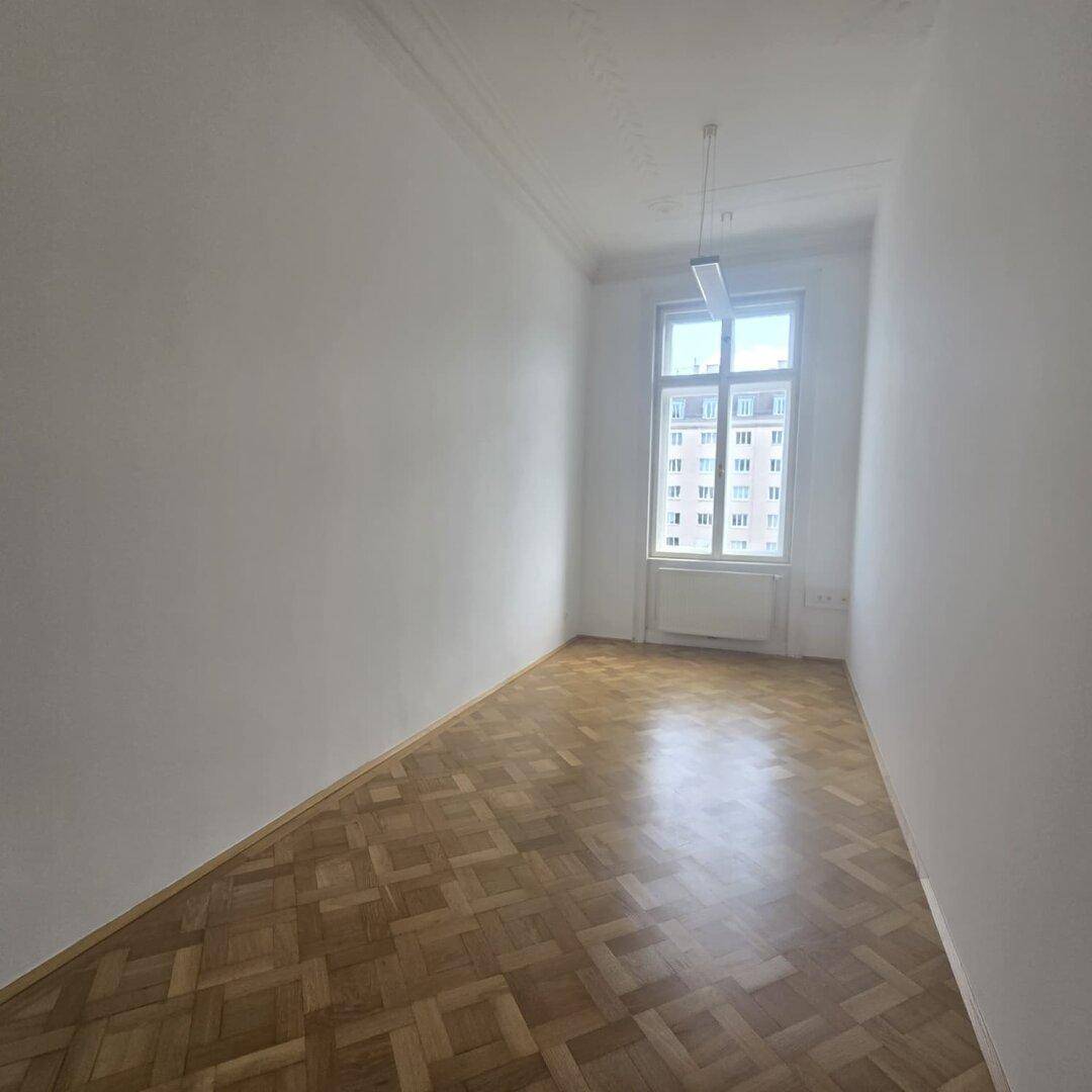 Schmaler Büroraum mit Fischgrätparkett, hohen Decken und einem großen Fenster, das den Raum erhellt.