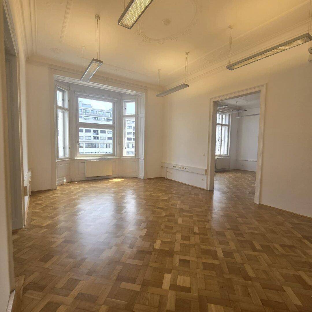 Geräumiger Büroraum mit Fischgrätparkett, hohen Decken und einem Erkerfenster, das viel Tageslicht hereinlässt.