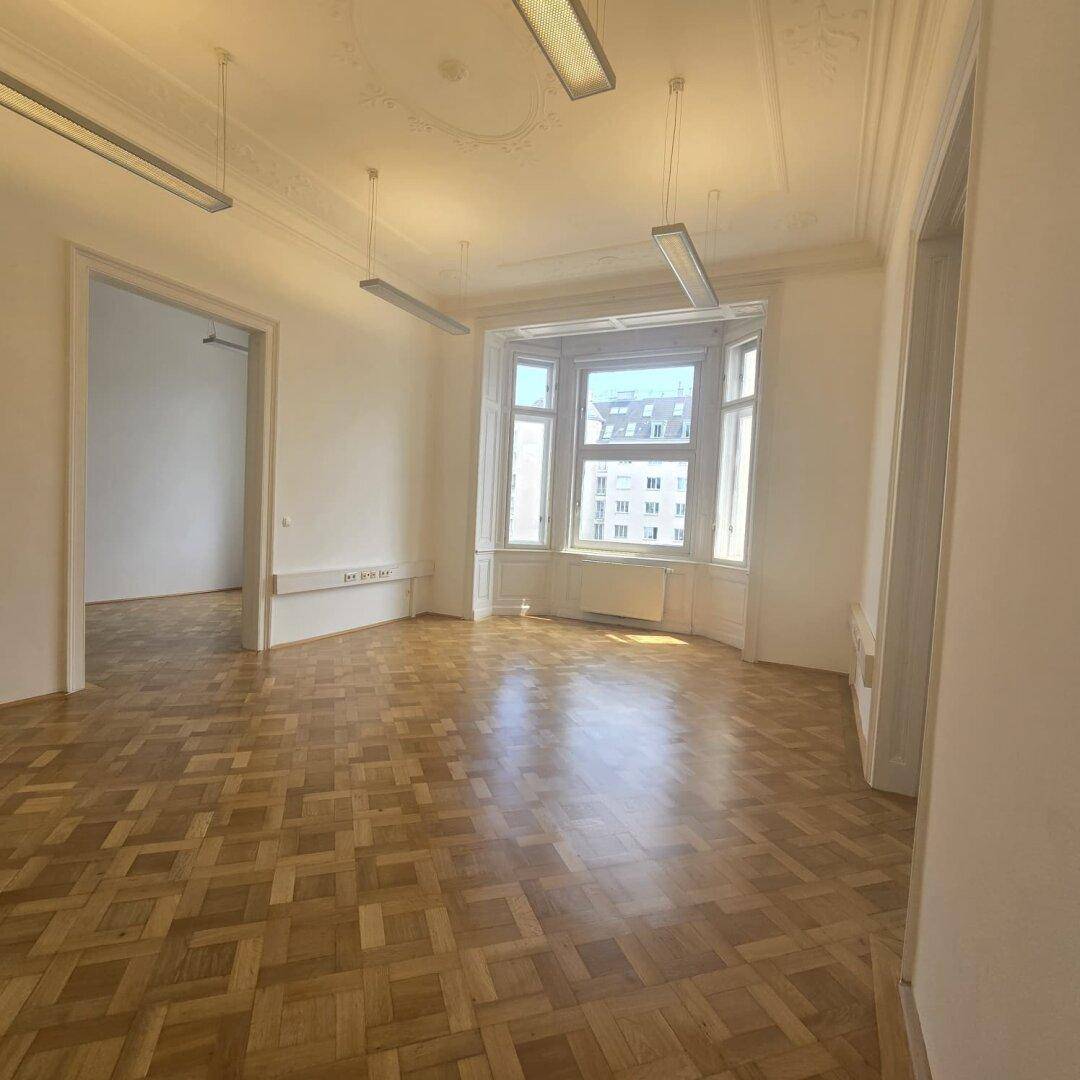 Großer Büroraum mit Fischgrätparkett, hohen Decken und einem Erkerfenster, das viel natürliches Licht bietet.