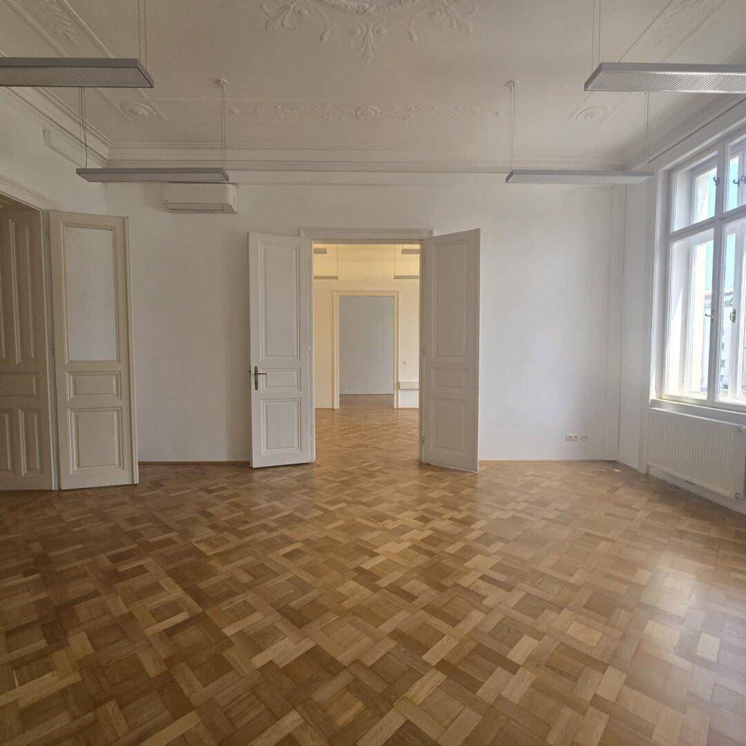 Großer Büroraum mit Fischgrätparkett, hohen Decken und zwei Fenstern, die den Raum mit Licht füllen.