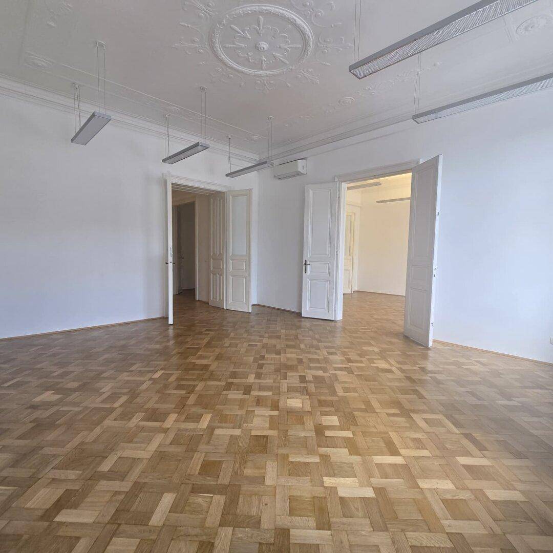 Heller Büroraum mit Fischgrätparkett, hohen Decken und mehreren Türen, die zu anderen Bereichen führen.