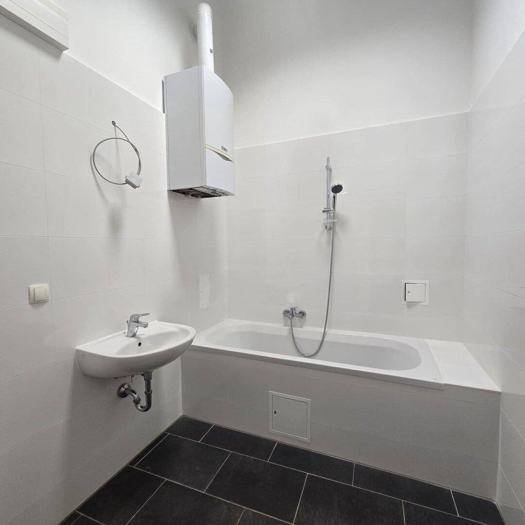 Badezimmer mit Badewanne, Waschbecken und weißen Fliesen, ergänzt durch einen Warmwasserbereiter.