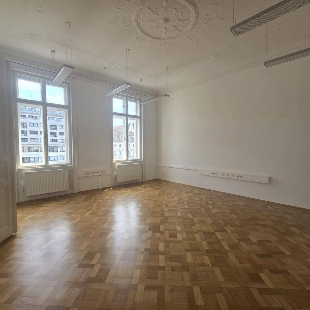 Heller Büroraum mit Fischgrätparkett, hohen Decken und zwei großen Fenstern, die viel Tageslicht hereinlassen.