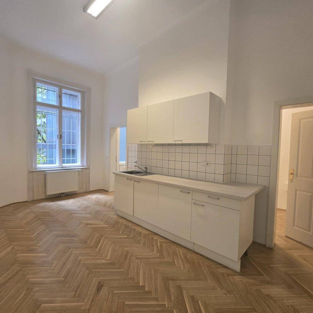 Einfache Küchenzeile mit weißen Schränken und Spüle in einem Raum mit Fischgrätparkett und Fenster.
