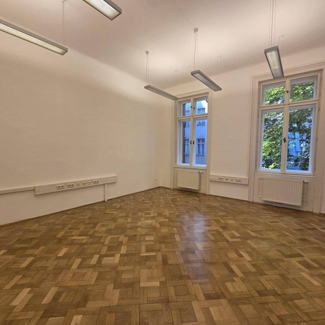 Geräumiger Büroraum mit Fischgrätparkett, hohen Decken und zwei Fenstern mit Blick ins Grüne.