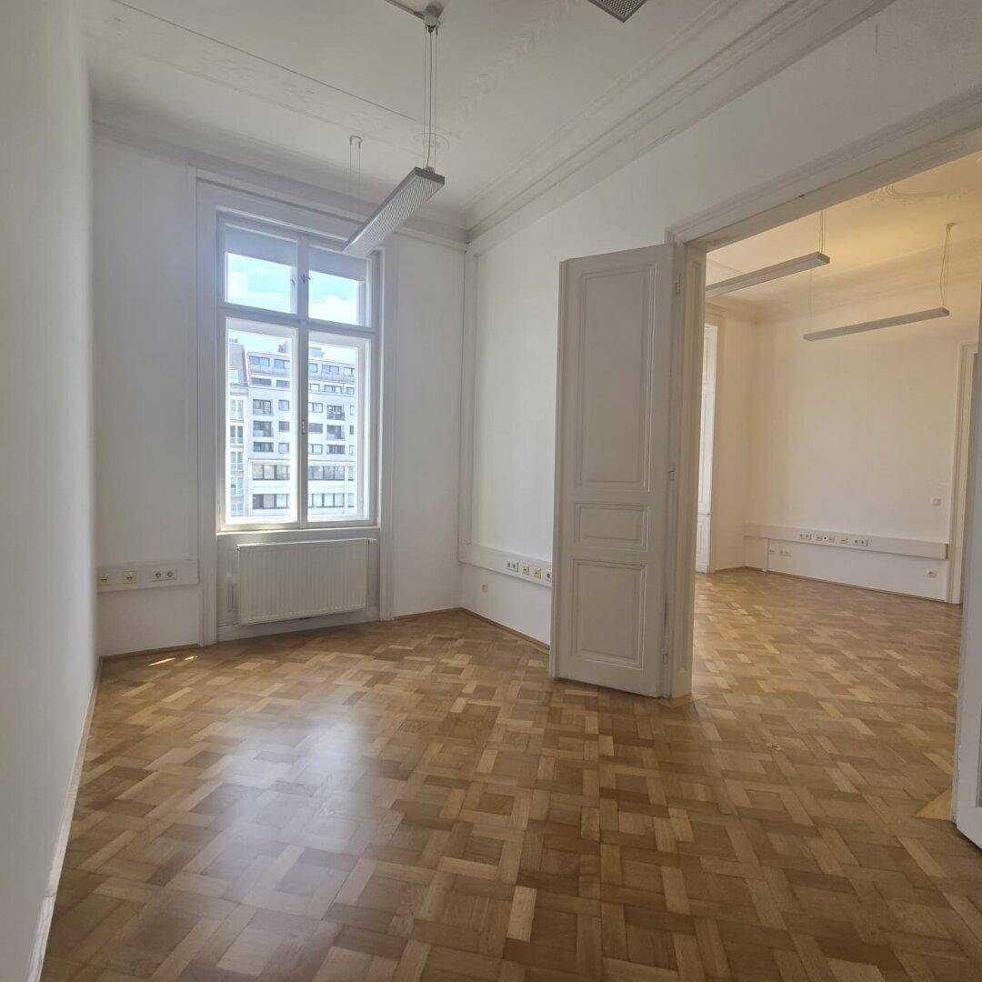 Leerer Büroraum mit Fischgrätparkett, hohen Decken und einem großen Fenster, das Tageslicht spendet.