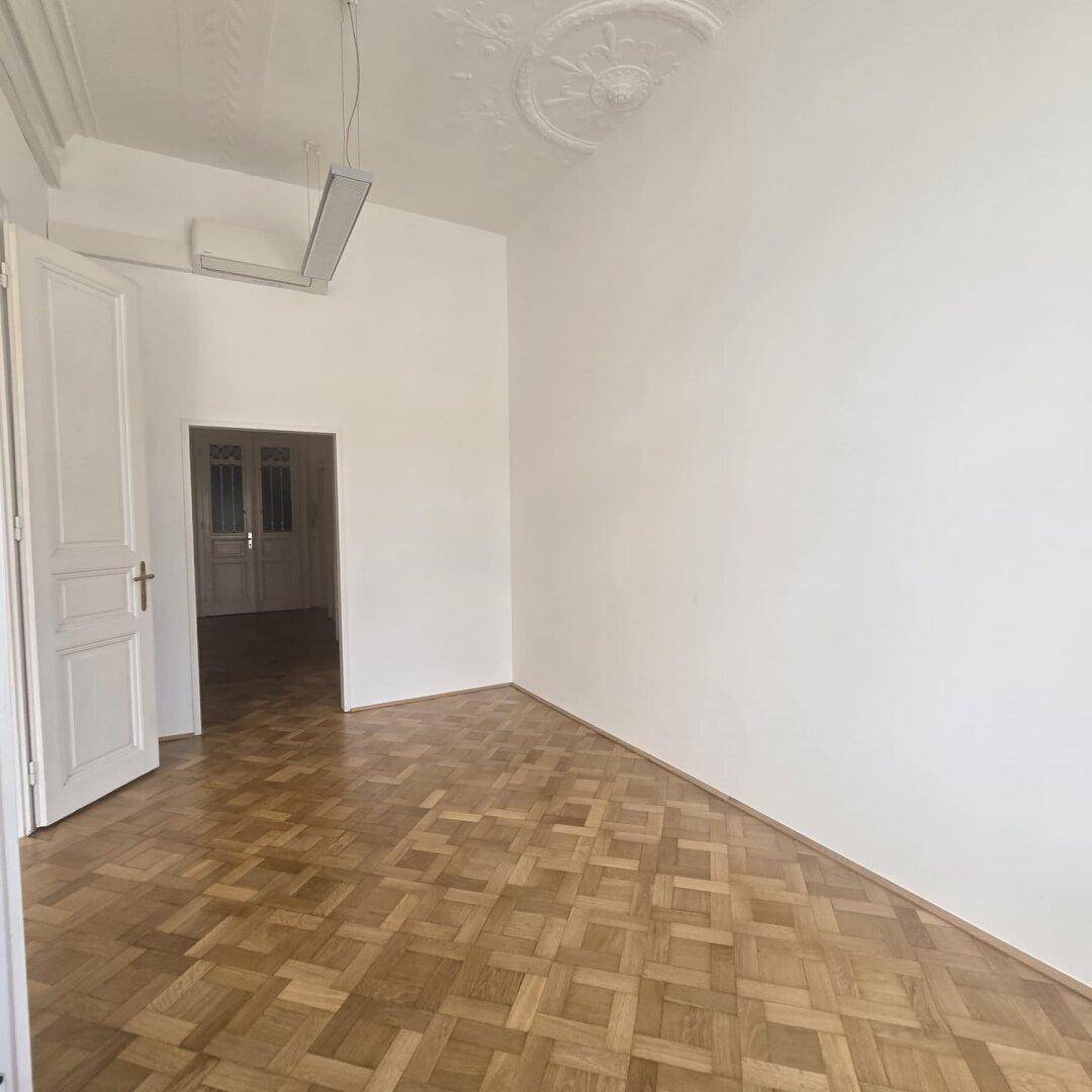 Leerer Büroraum mit Parkettboden und einer Tür, die in einen weiteren Bereich führt.