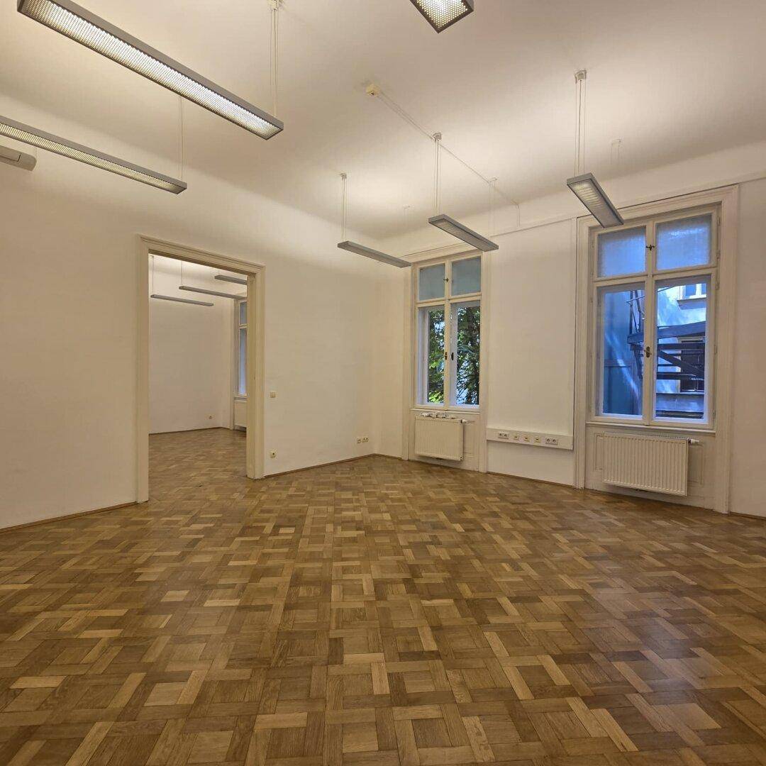 Großer Büroraum mit zwei Fenstern und Parkettboden, der viel Tageslicht hereinlässt.