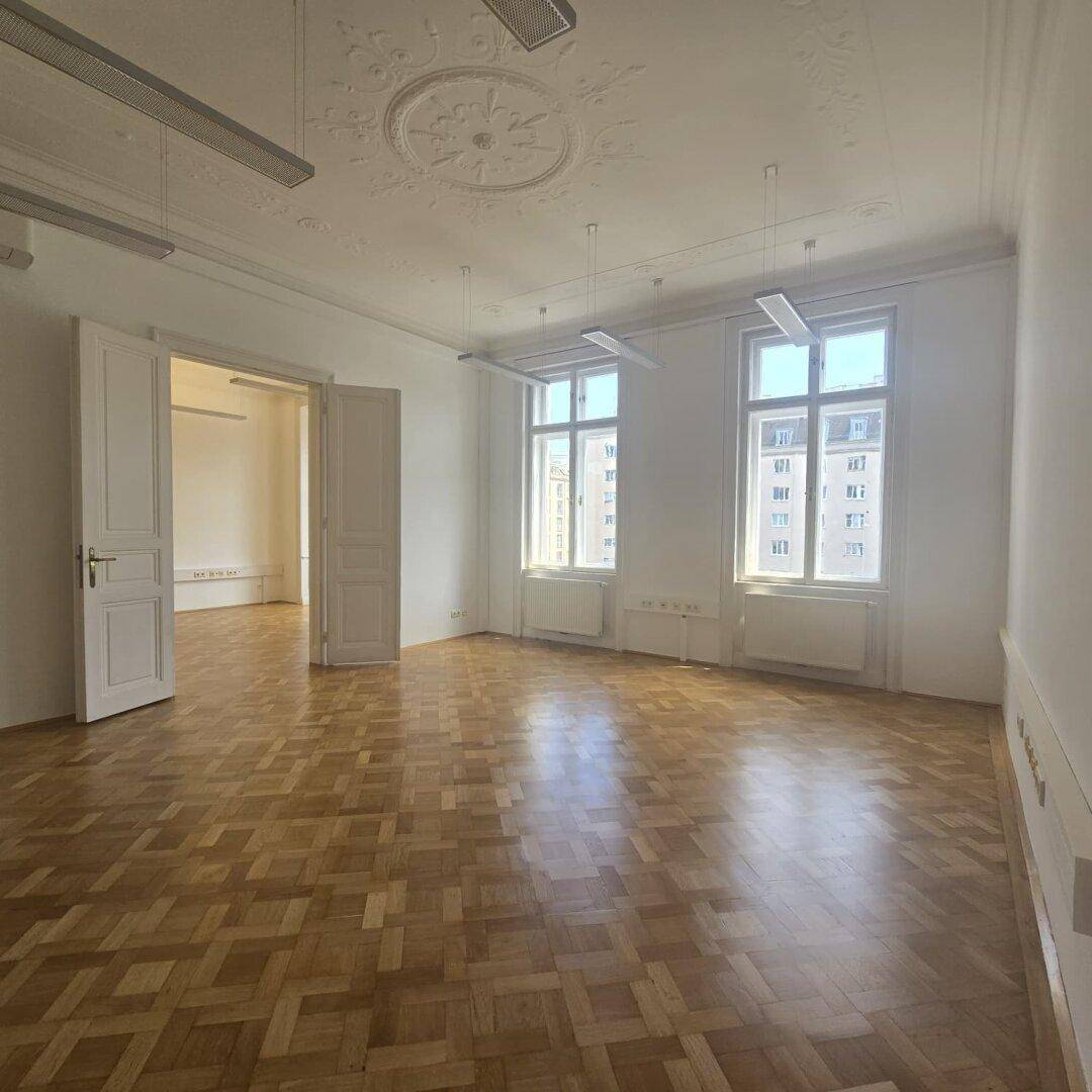 Heller Büroraum mit klassischem Fischgrätparkett, hohen Decken und zwei großen Fenstern mit Stadtblick.