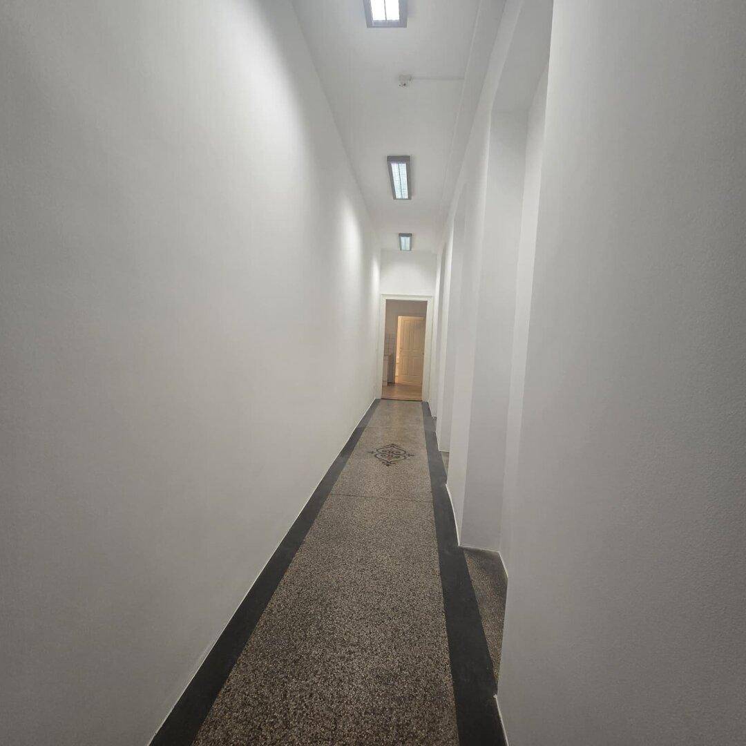 Langer, schmaler Flur mit Terrazzoboden und Deckenbeleuchtung, der zu weiteren Räumen führt.