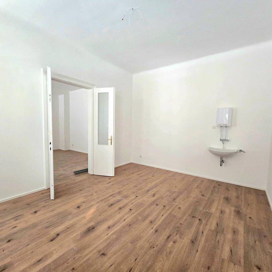 Heller Raum mit Holzboden, Waschbecken und Zugang zu weiteren Zimmern durch eine offene Tür.