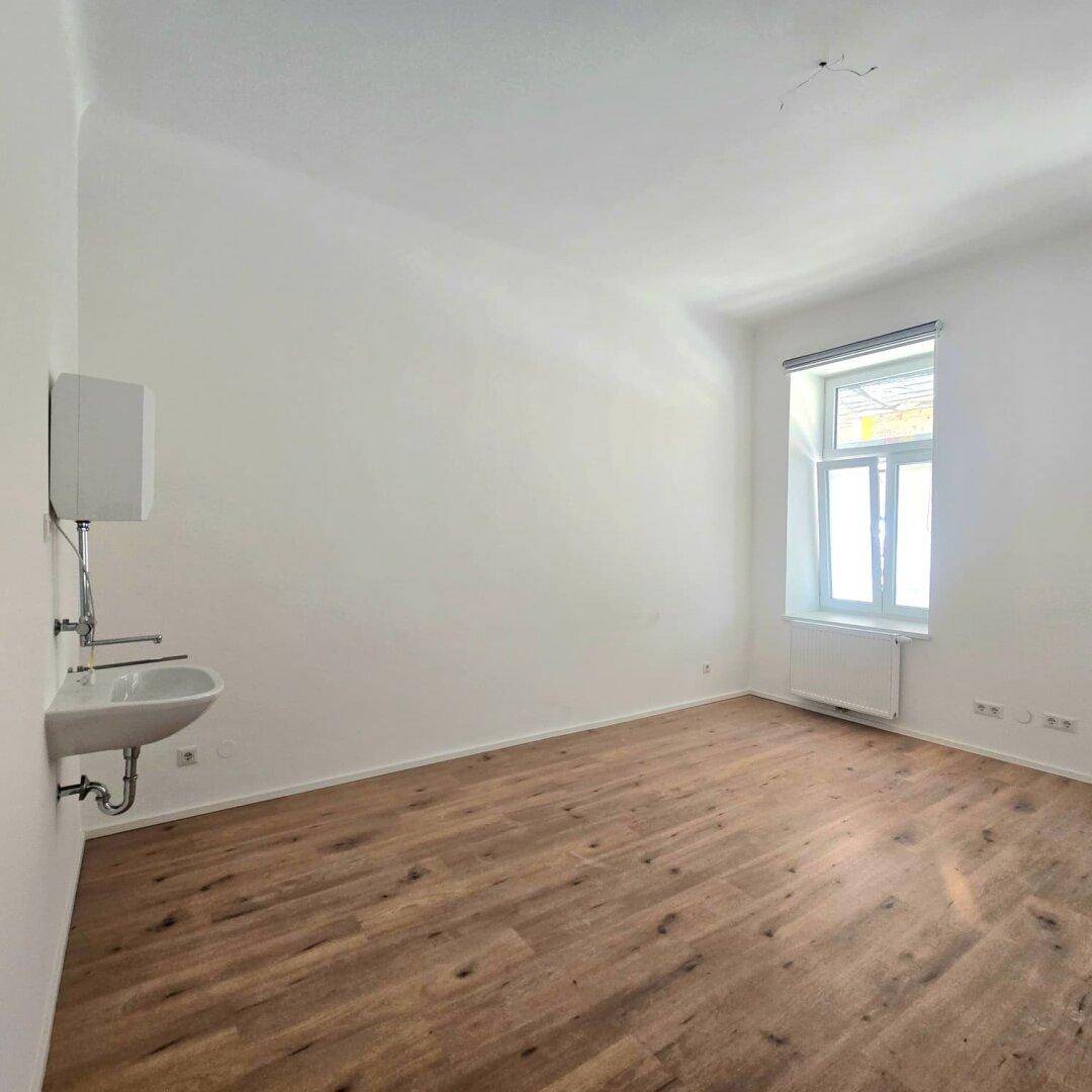 Funktionaler Raum mit Holzboden, Fenster und einem Waschbecken mit Warmwasserbereiter an der Wand.