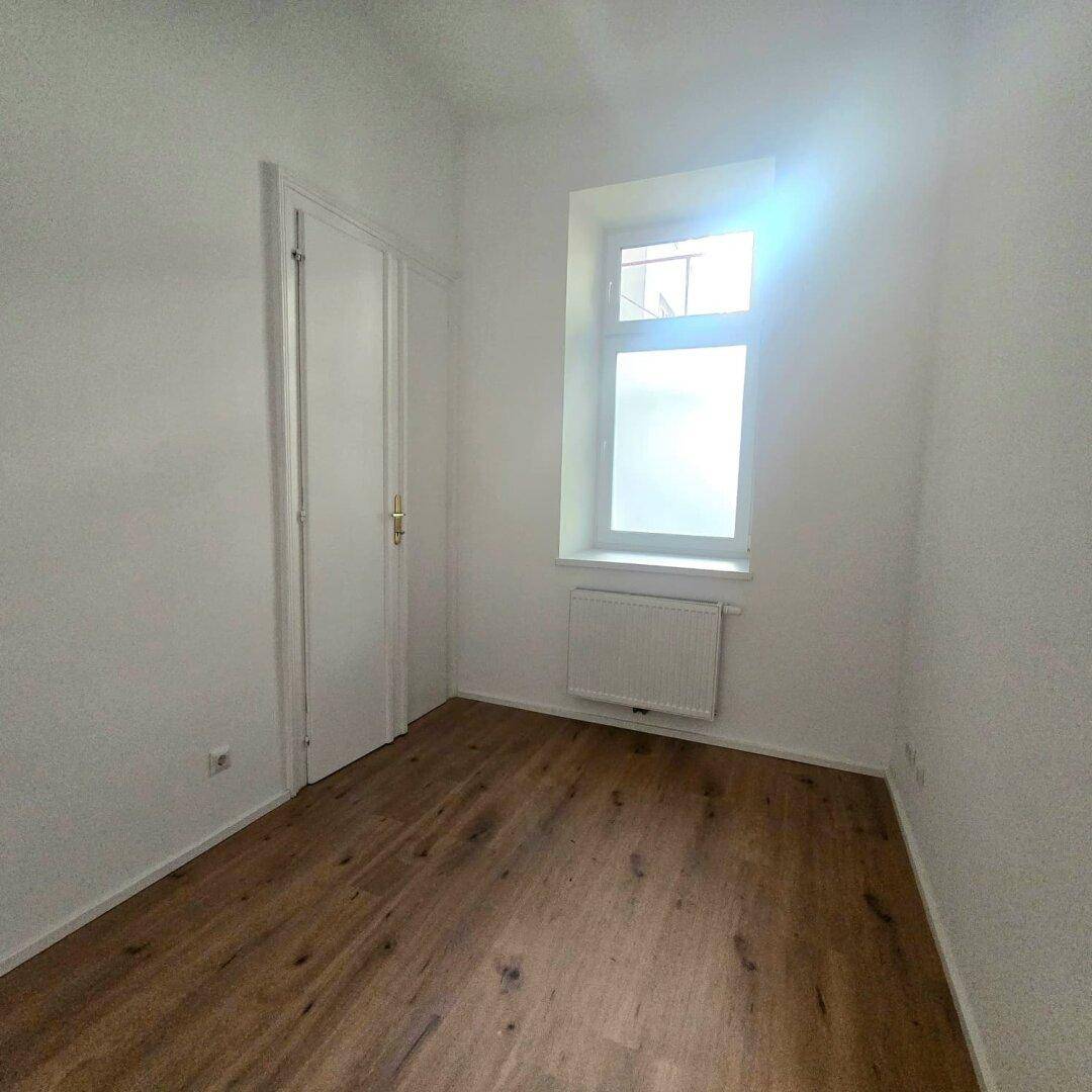 Kleines, helles Zimmer mit Holzboden, weißen Wänden, einem Fenster und einer geschlossenen Tür.