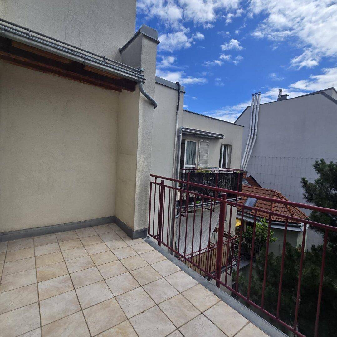 Heller Balkon mit Fliesenboden und rotem Geländer, umgeben von angrenzenden Gebäuden unter blauem Himmel.
