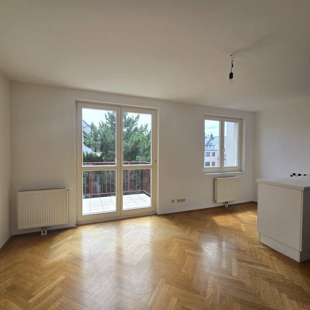 Helles Zimmer mit Fischgrätparkettboden, großen Fenstern und Zugang zu einem Balkon mit Blick ins Grüne.