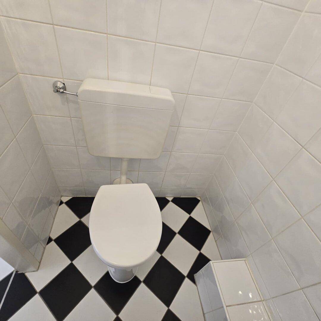 Separates WC mit weißen Fliesenwänden und einem schwarz-weißem Schachbrettmuster-Fliesenboden.