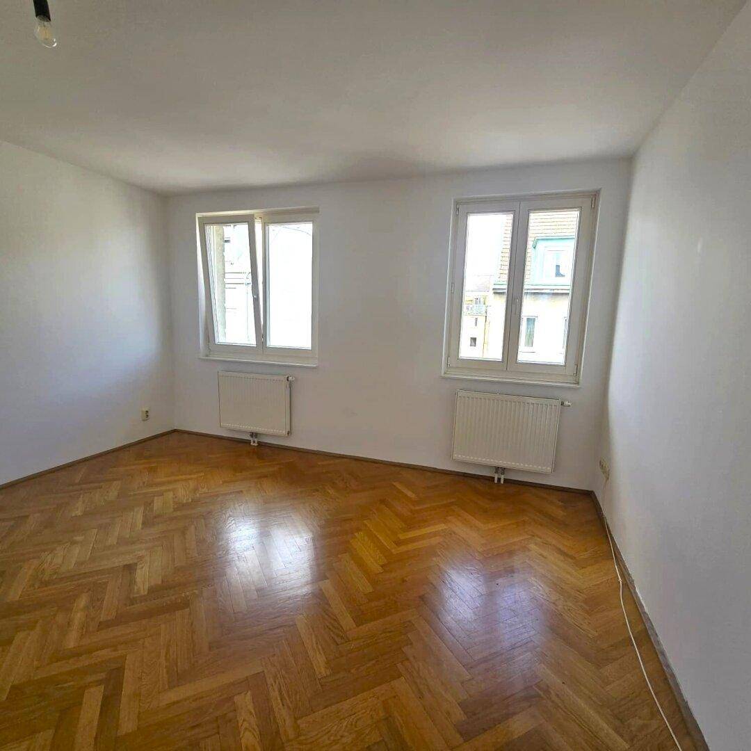 Geräumiges Zimmer mit Fischgrätparkettboden und zwei Fenstern, die für ausreichend Tageslicht sorgen.