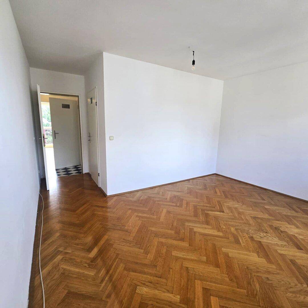 Helles Zimmer mit Fischgrätparkettboden und Zugang zu einem Flur mit Schachbrettmuster-Fliesen.