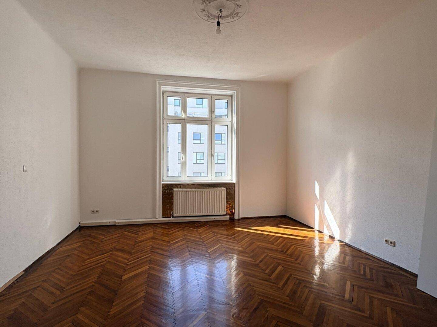 Geräumiges Zimmer mit Fischgrätparkett, Stuckdecke und einem großen Fenster.
