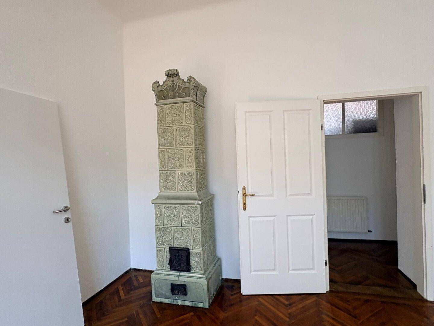 Zimmer mit traditionellem Kachelofen, Fischgrätparkett und weiß gestrichenen Wänden.