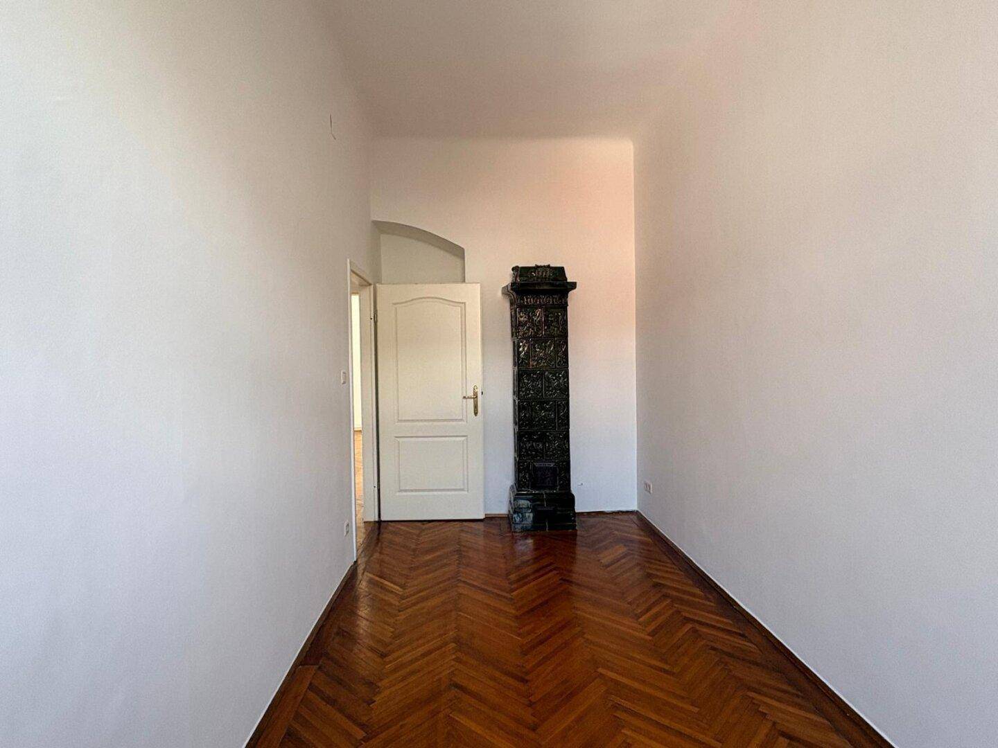 Zimmer mit dunklem Kachelofen, Fischgrätparkett und weiß gestrichenen Wänden.