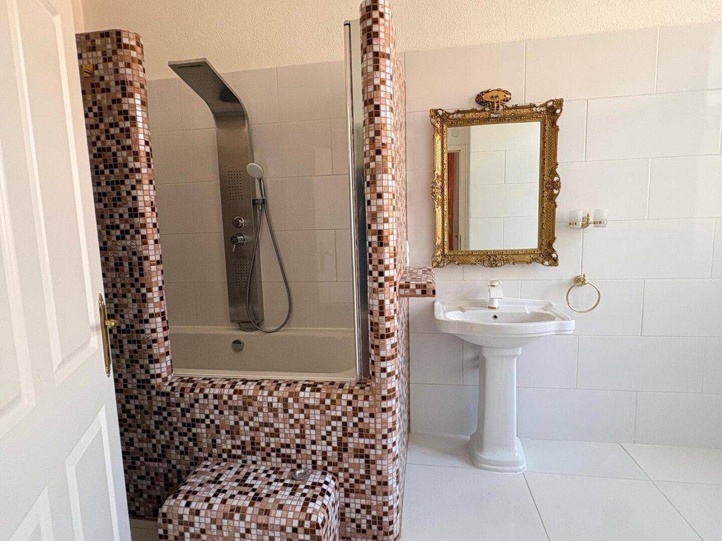 Badezimmer mit Mosaikfliesen an der Badewanne, Duscharmatur und Standwaschbecken.