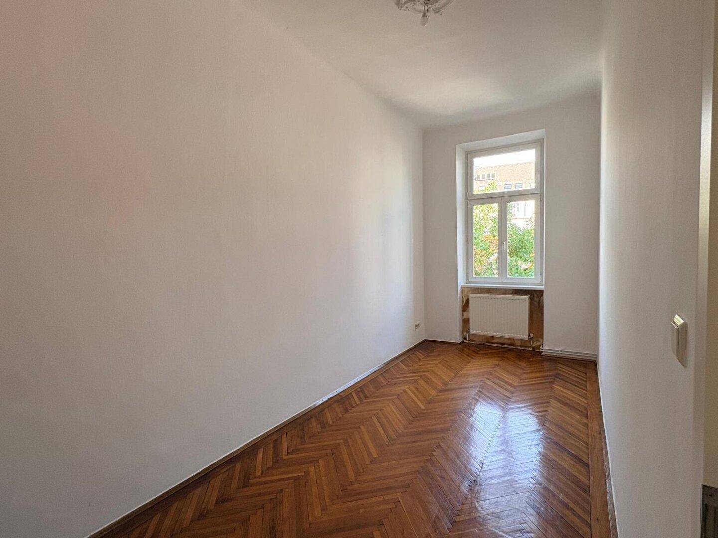Helles Schlafzimmer mit Fischgrätparkett und Blick ins Grüne durch das Fenster.