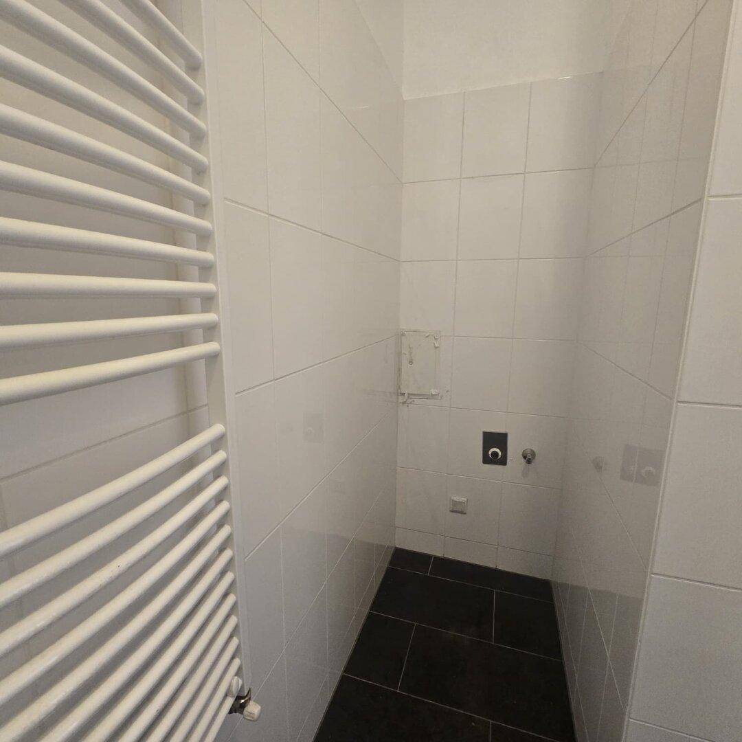 Bereich im Badezimmer mit weißen Fliesen und dunklem Boden, vorbereitet für eine Waschmaschine.