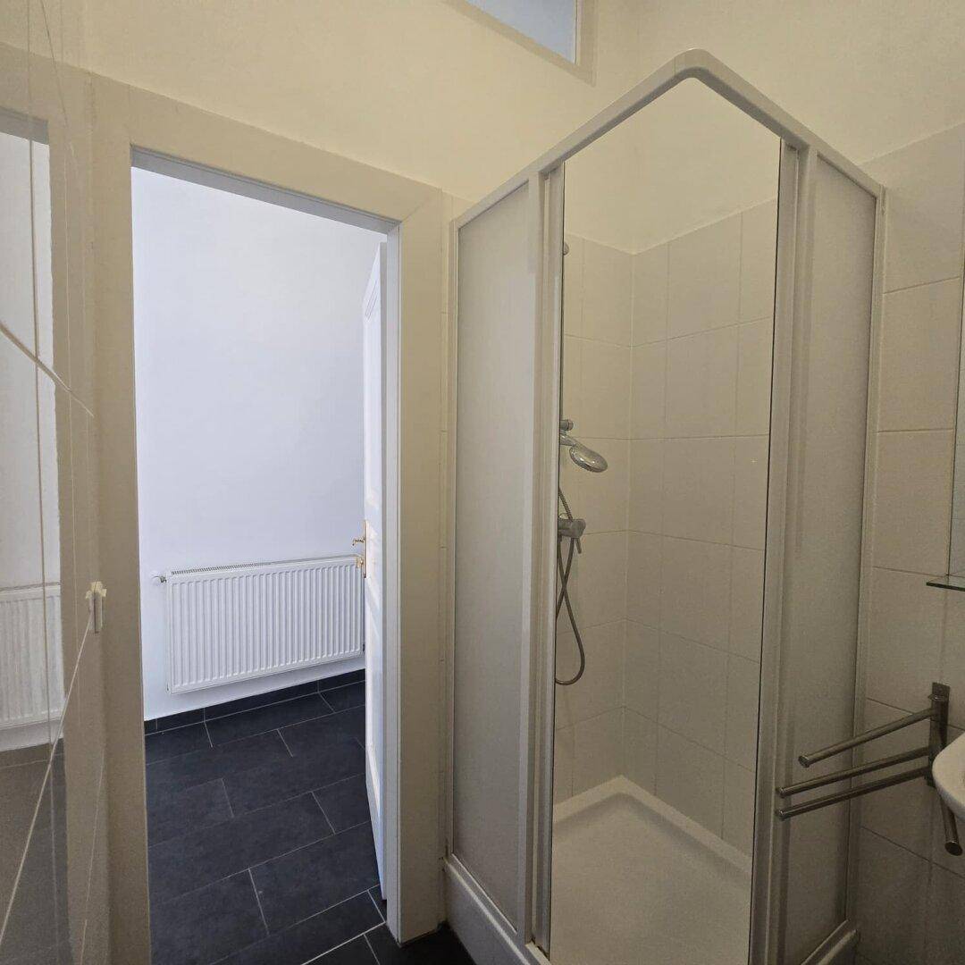 Badezimmer mit Duschkabine und Zugang zu einem weiteren Raum mit Heizkörper.