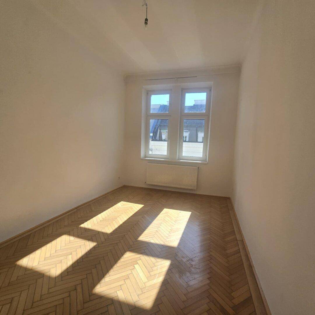 Freundliches Zimmer mit Fischgrätparkett und zwei Fenstern, die den Raum mit Sonnenlicht durchfluten.