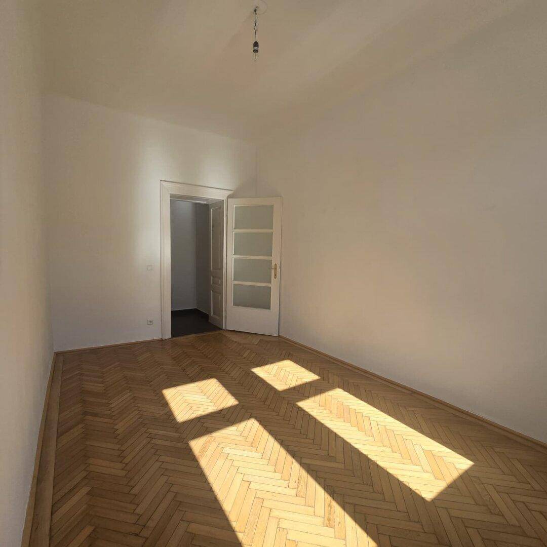 Geräumiges Zimmer mit Fischgrätparkett und einer Tür mit Glaseinsätzen, die den Raum erhellt.