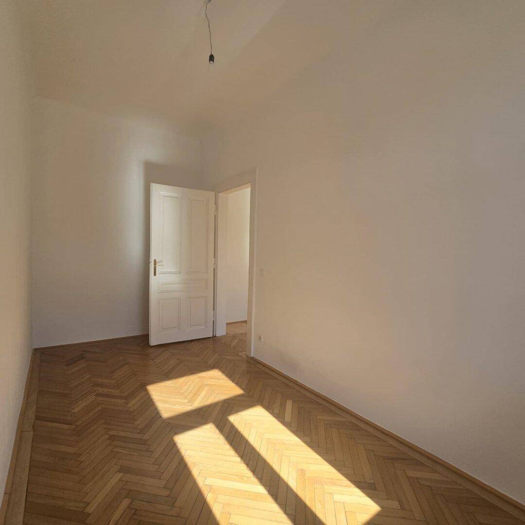 Sonniger Raum mit Fischgrätparkett und einer weißen Tür, die den Zugang zu einem anderen Bereich ermöglicht.