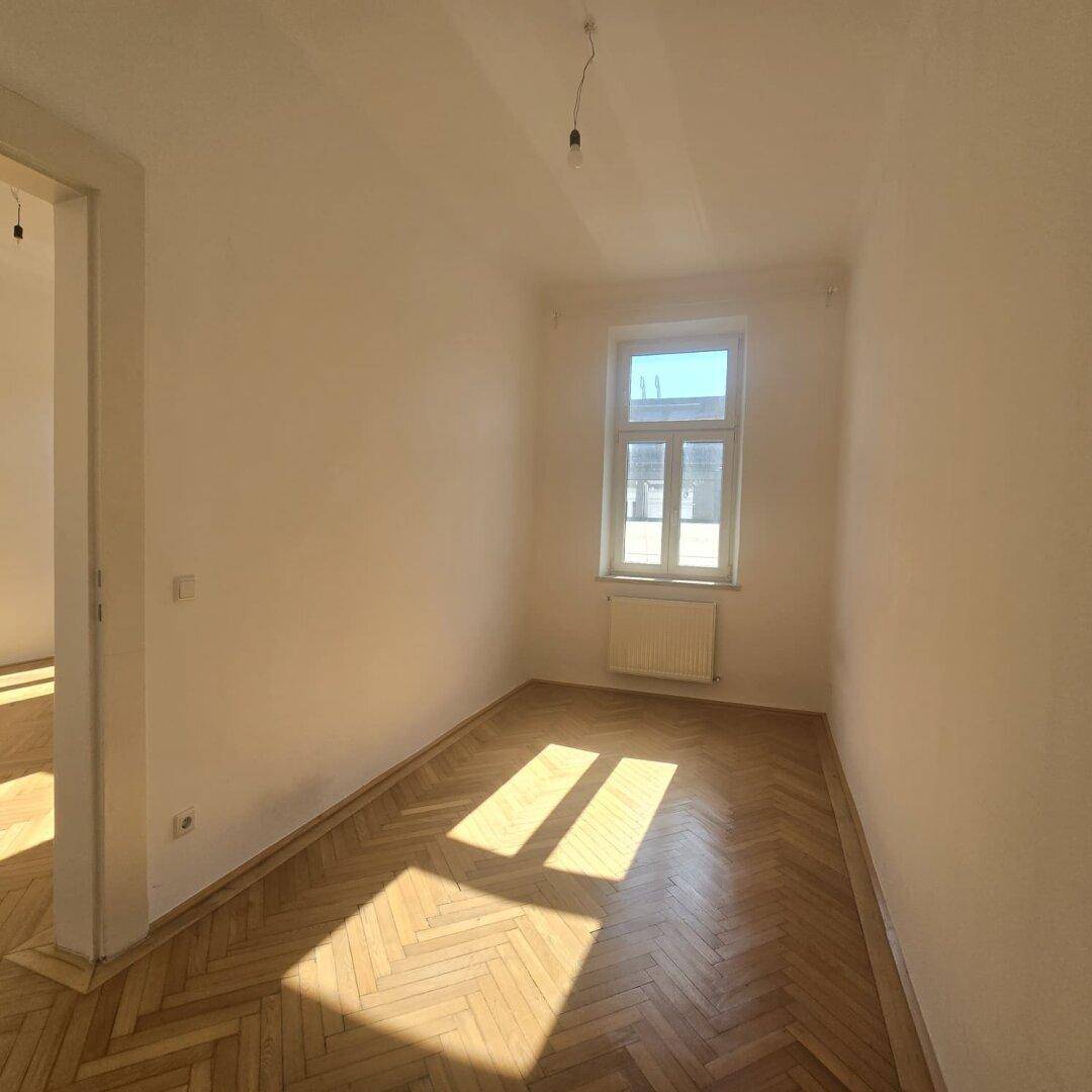 Helles, schmales Zimmer mit Fischgrätparkett und einem Fenster, das viel Tageslicht hereinlässt.
