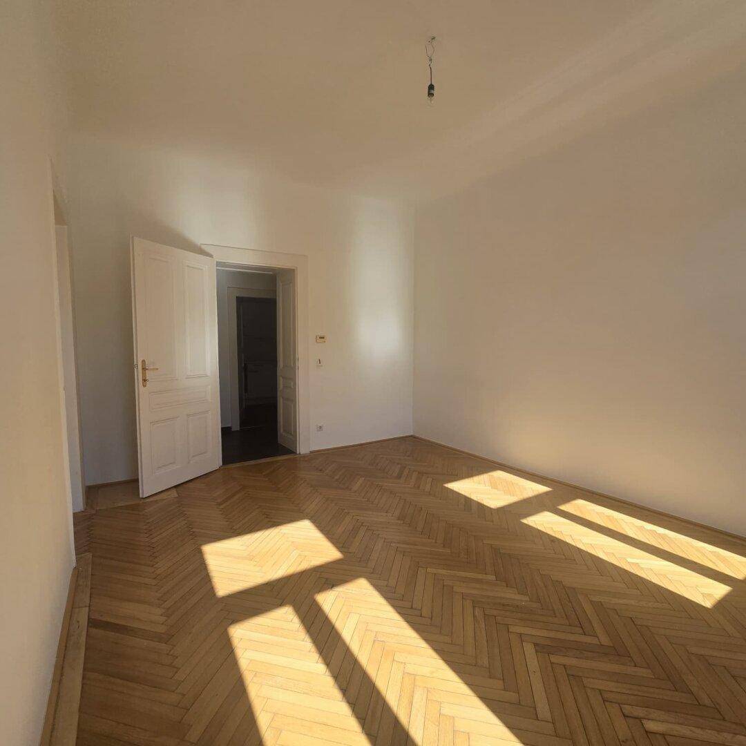 Geräumiges Zimmer mit Fischgrätparkett und einer geöffneten Tür, die in einen weiteren Raum führt.
