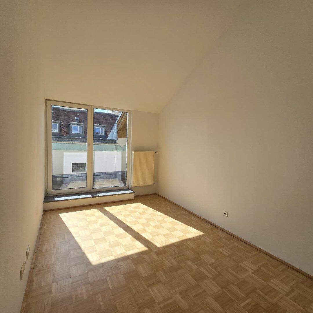 Heller Raum mit Parkettboden, weißen Wänden und großem Fenster, das viel Sonnenlicht hereinlässt.