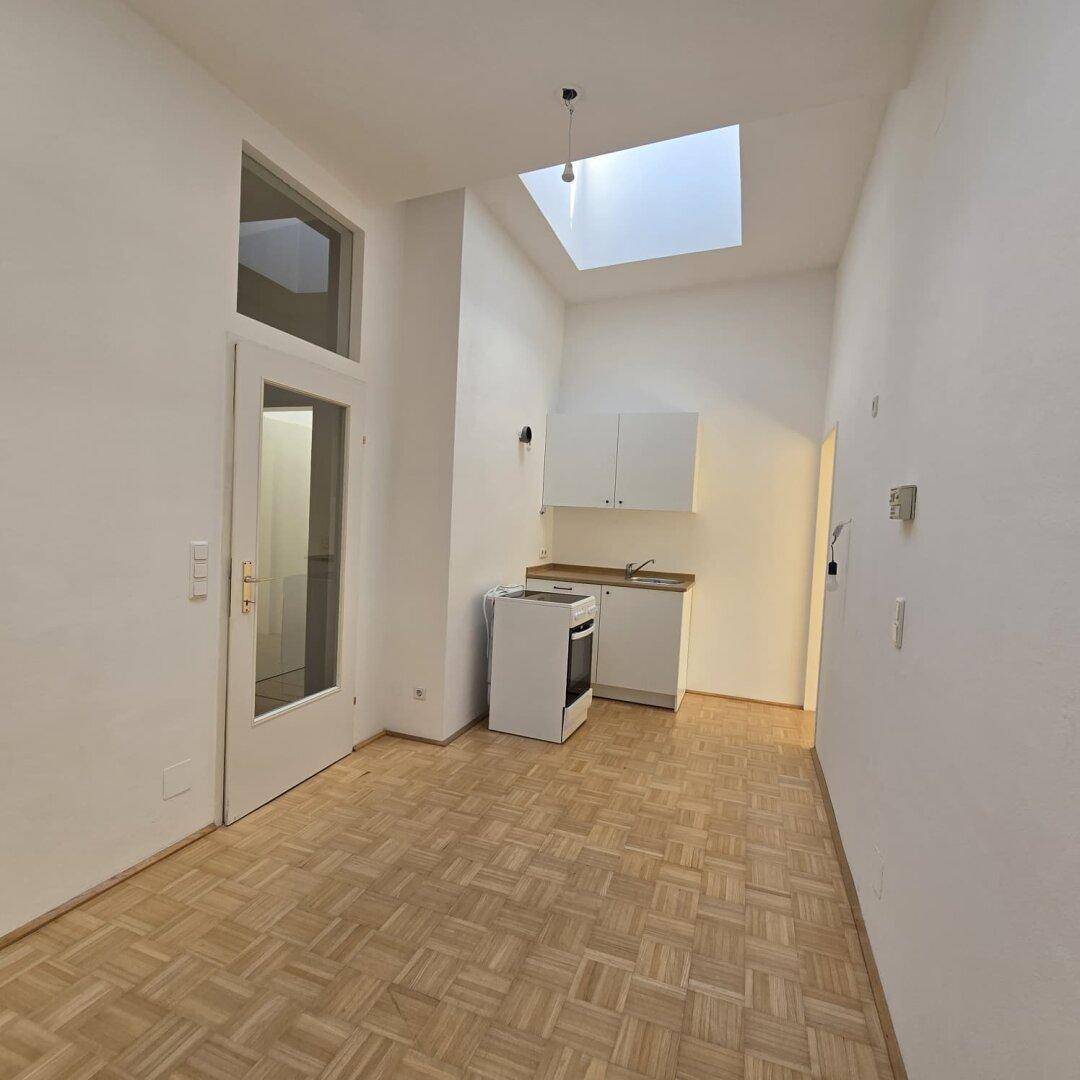 Heller Raum mit kleiner Küchenzeile, Parkettboden und einem Oberlicht, das Tageslicht spendet.