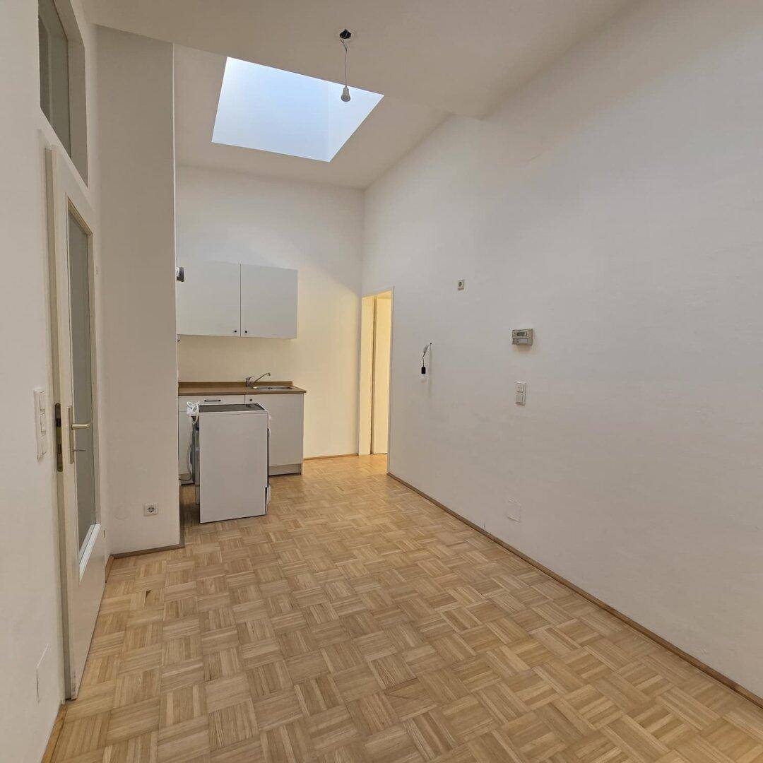 Geräumiger Raum mit Küchenzeile, Parkettboden und hoher Decke, beleuchtet durch ein Oberlicht.