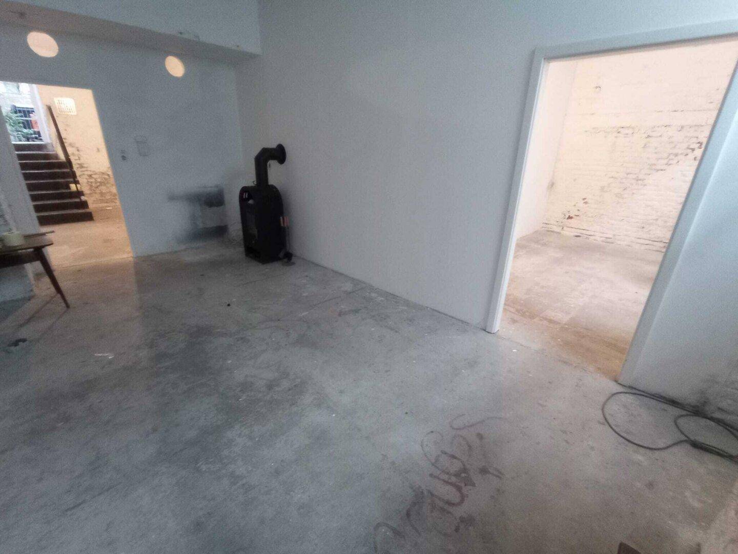 Kellerraum mit Betonboden, einem Ofen und Blick auf eine Treppe zu einem weiteren Bereich, der renoviert werden muss.