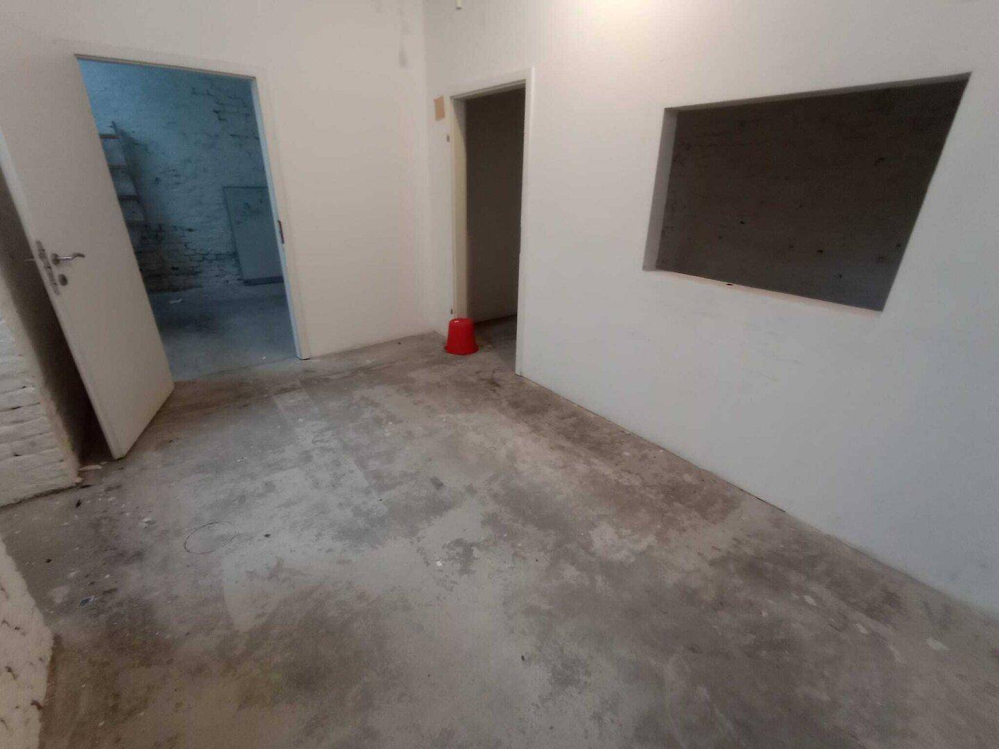 Kellerraum mit Betonboden, weißen Wänden und einem Durchgang zu einem weiteren Raum, der noch unfertig ist.