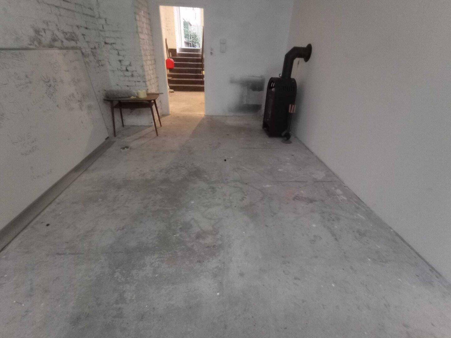 Kellerraum mit Betonboden, einem Ofen und einer Treppe im Hintergrund, die zu einem anderen Bereich führt.