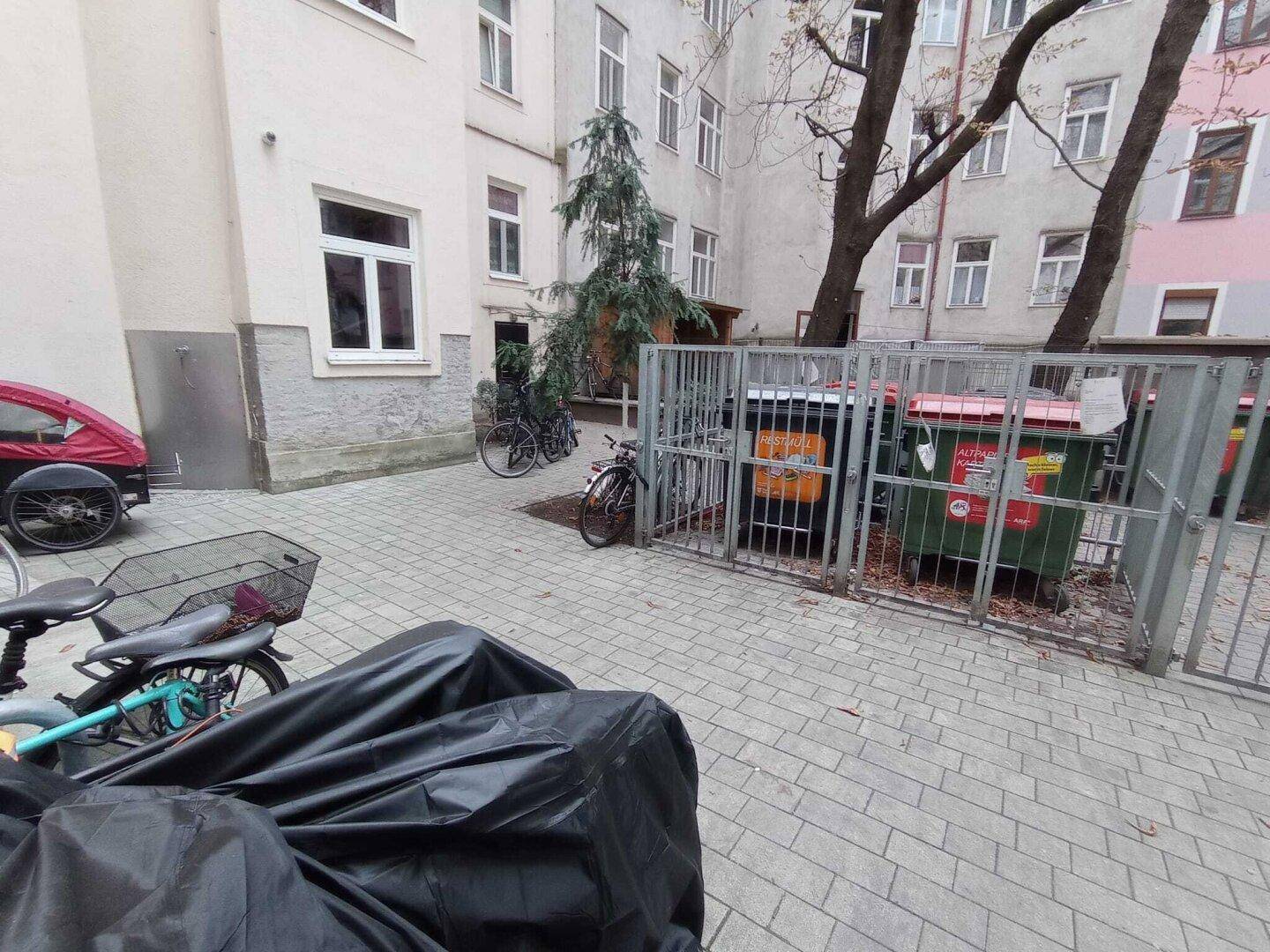 Innenhof mit Mülltonnen in einem Gitterkäfig, Fahrrädern und einem Mehrfamilienhaus im Hintergrund.
