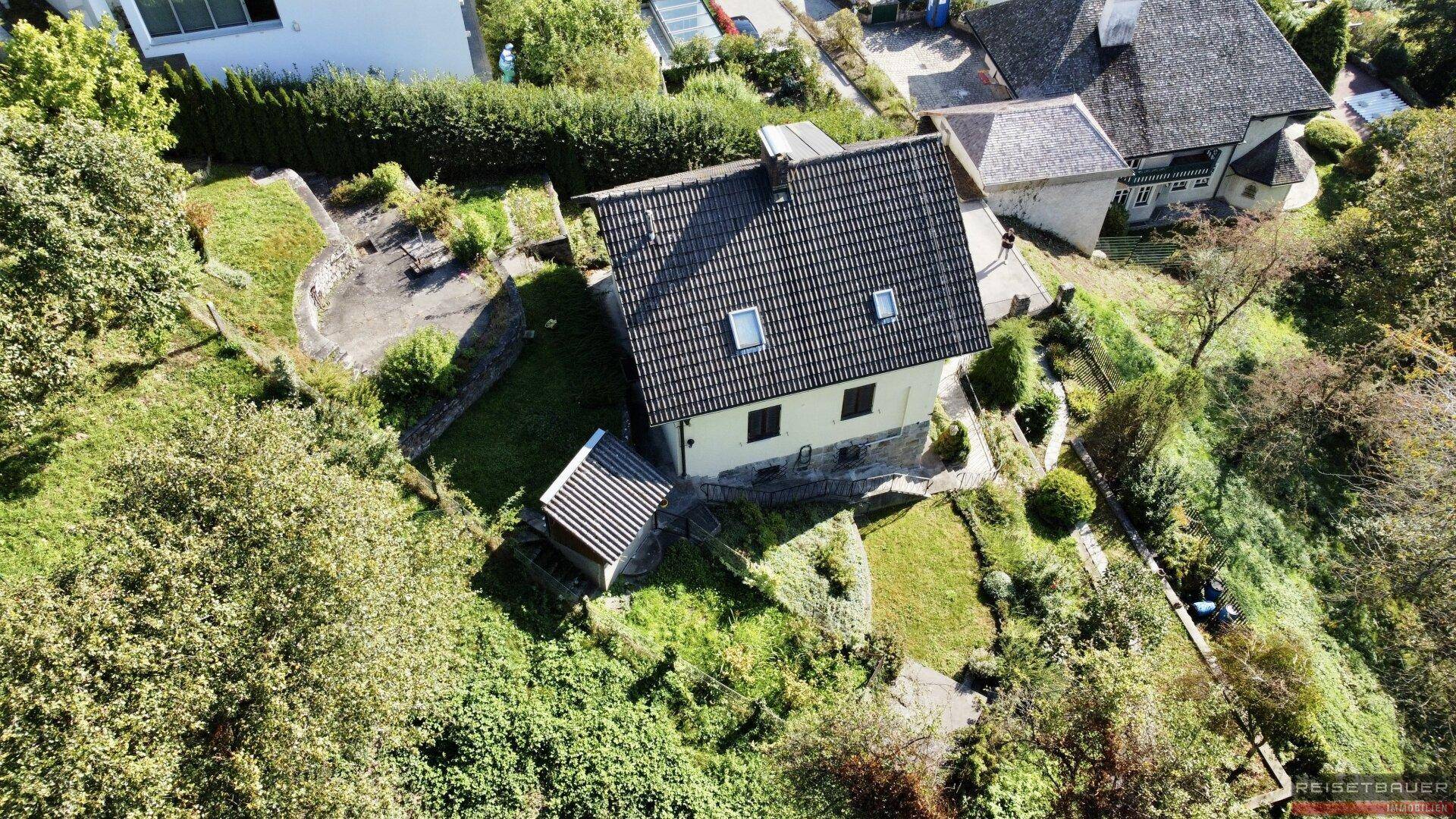 Luftaufnahme eines Hauses mit Garten und Nebengebäude in einer grünen, hügeligen Umgebung mit Bäumen.