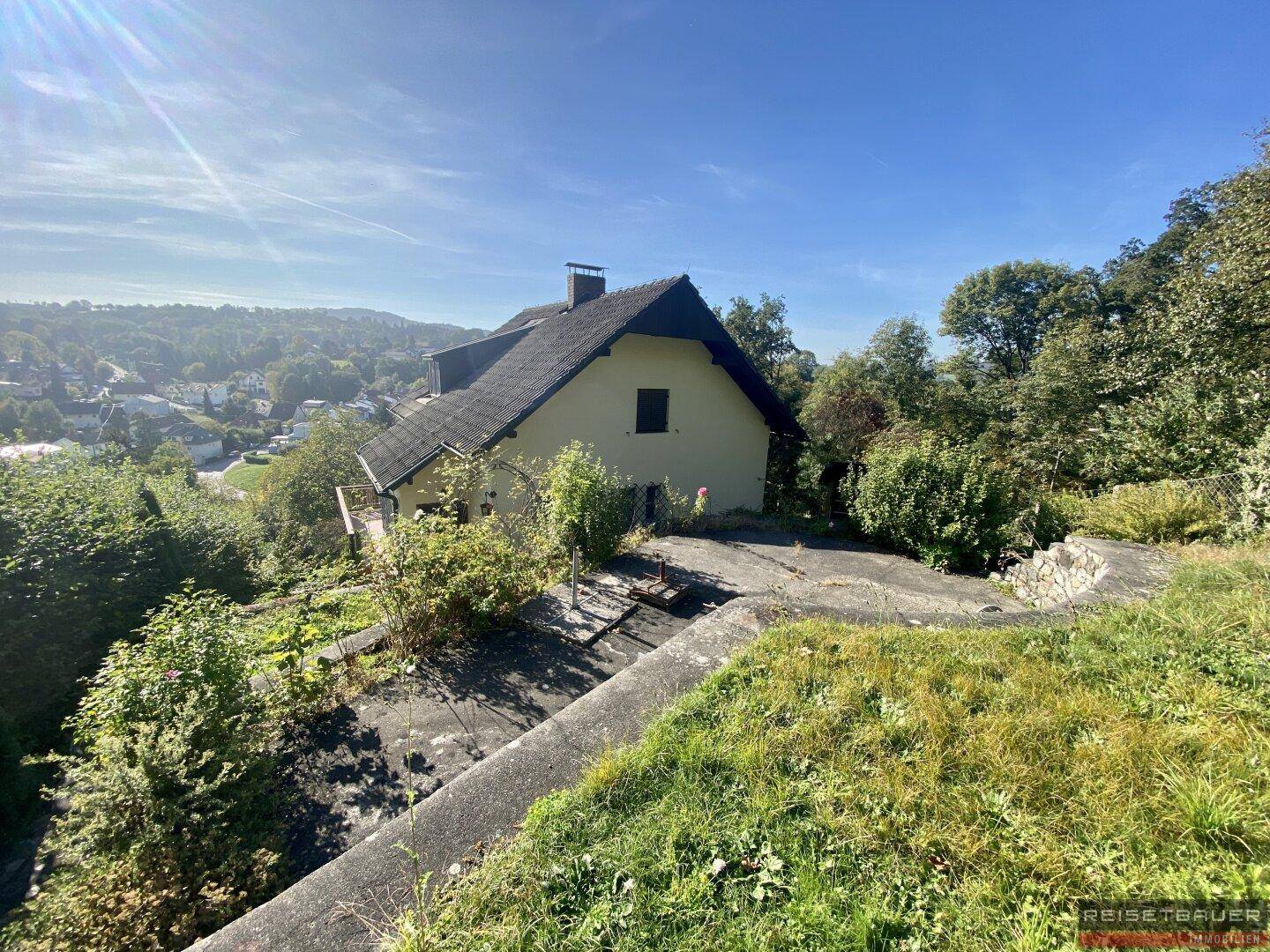 Haus in Hanglage mit Blick auf die umliegende grüne Landschaft unter blauem Himmel und Sonnenschein.