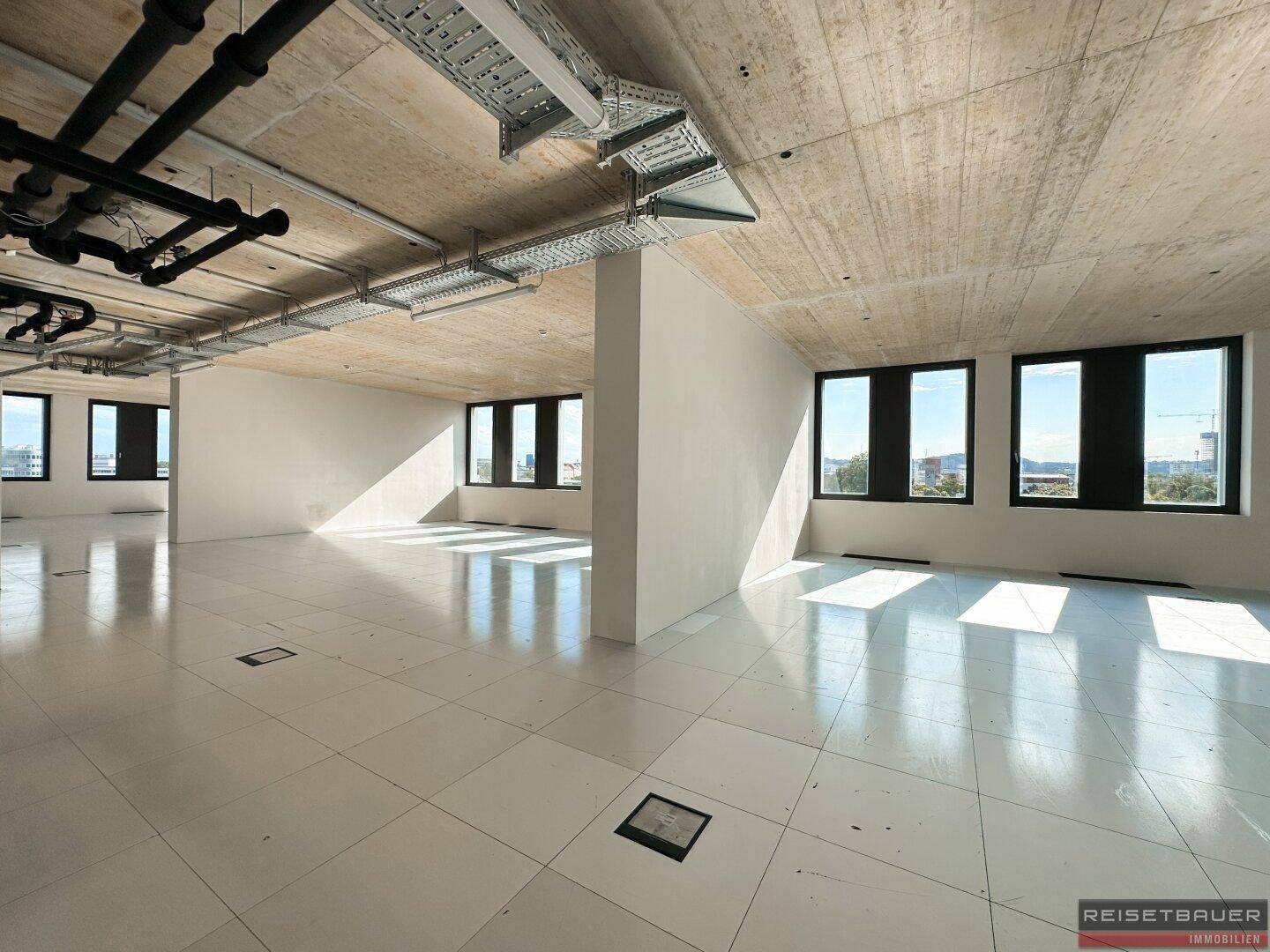 Moderner Büroraum im Rohbau mit sichtbaren Installationen und großen Fenstern für viel Tageslicht.