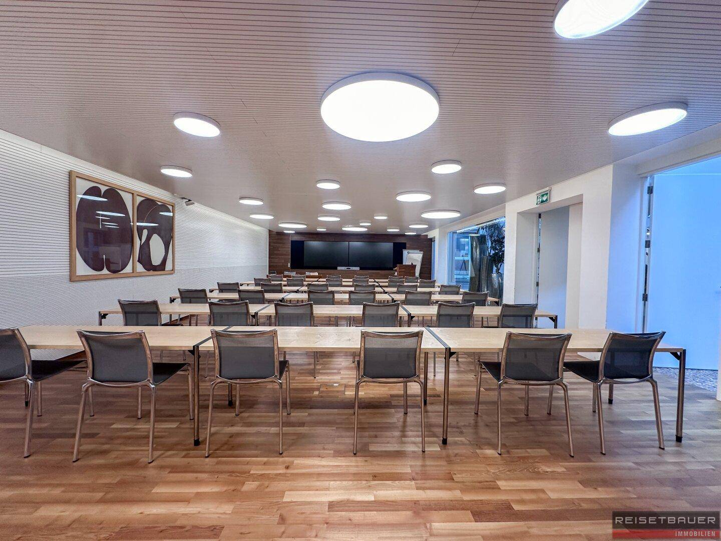 Großer Konferenzraum mit Reihenbestuhlung, Holzboden und moderner Deckenbeleuchtung.