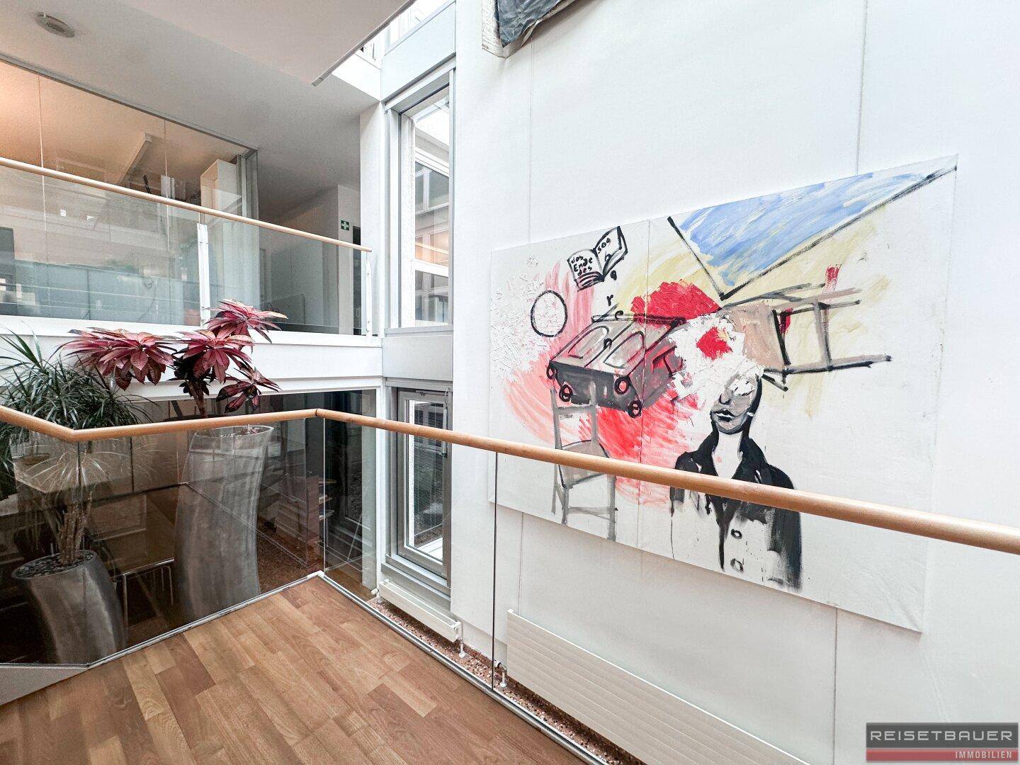 Heller Flur mit Kunstwerk, Holzgeländer und Blick auf eine moderne Glasfassade.