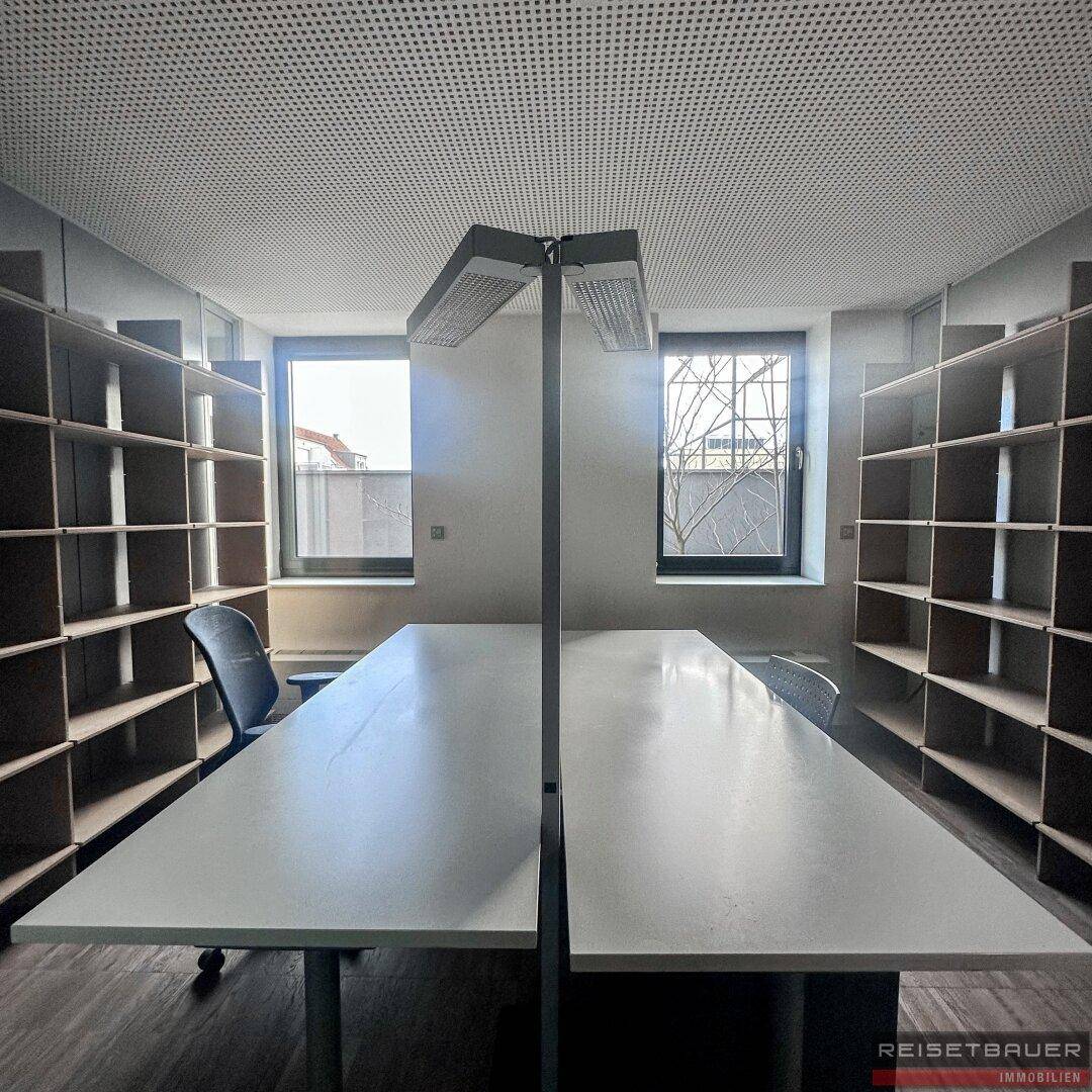 Heller Büroraum mit zwei Arbeitsplätzen, großen Fenstern und Regalen an den Wänden.