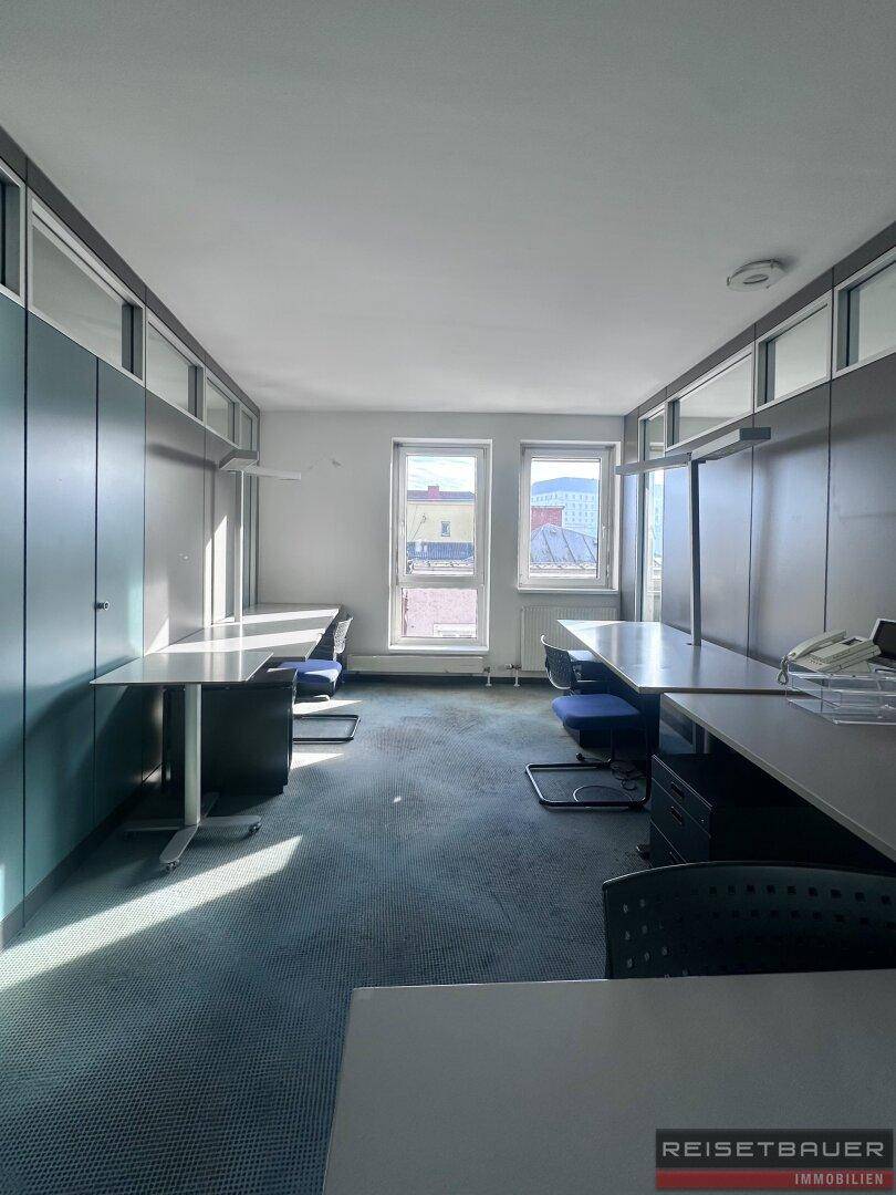 Helles Büro mit Teppichboden, modularen Arbeitsplätzen und großen Fenstern.